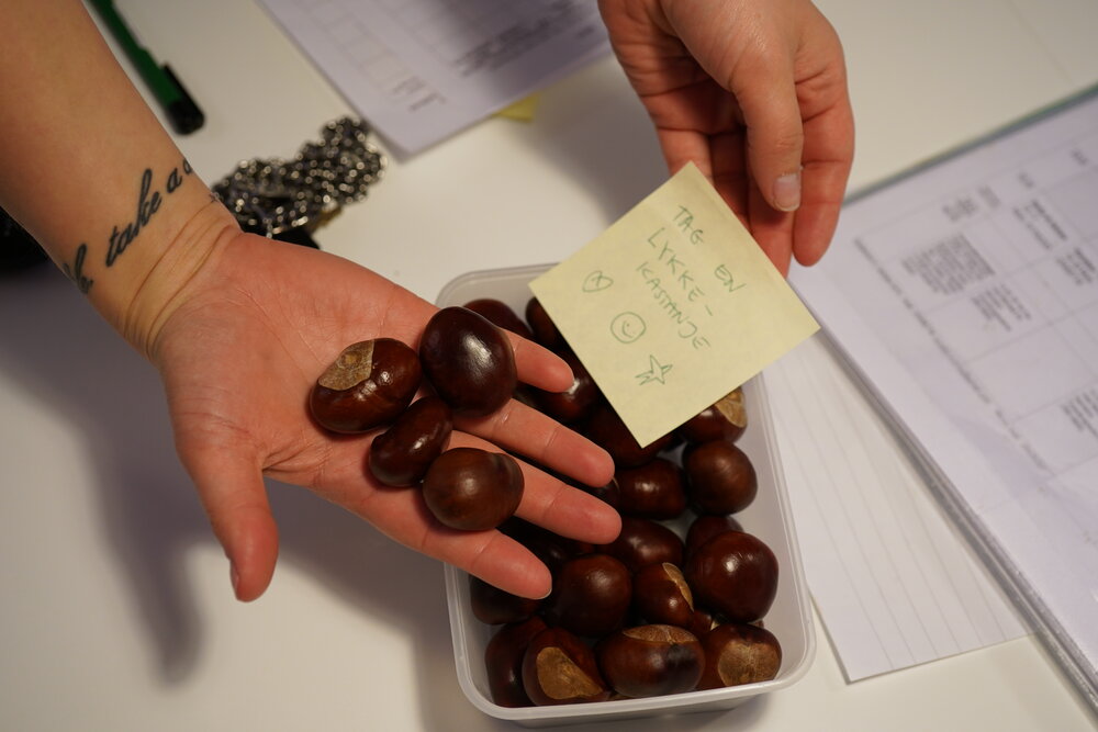 A caregiver holding a handful of chestnuts above a tray of chestnuts, together with a note "take a lucky chestnut" in Danish
