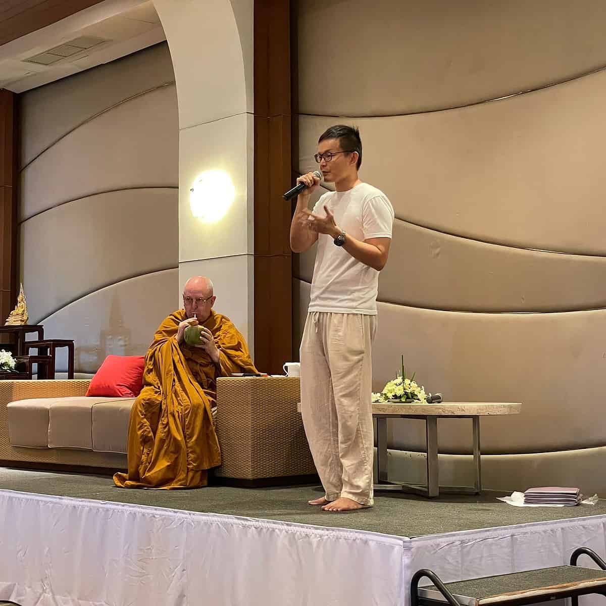 Man holding microphone on a stage, next to a monk on a sofa drinking from a coconut.